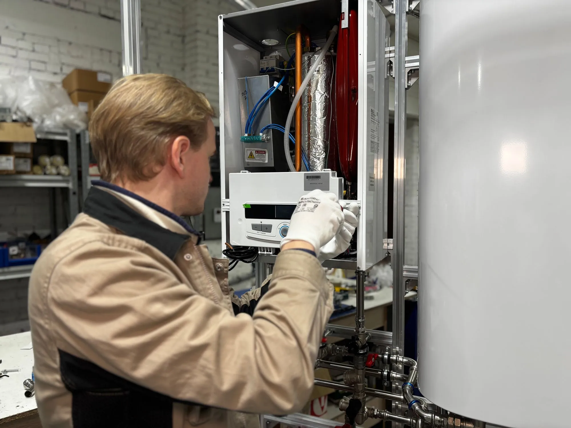 A heating technician in work gloves servicing the open front of a wall-mounted gas boiler