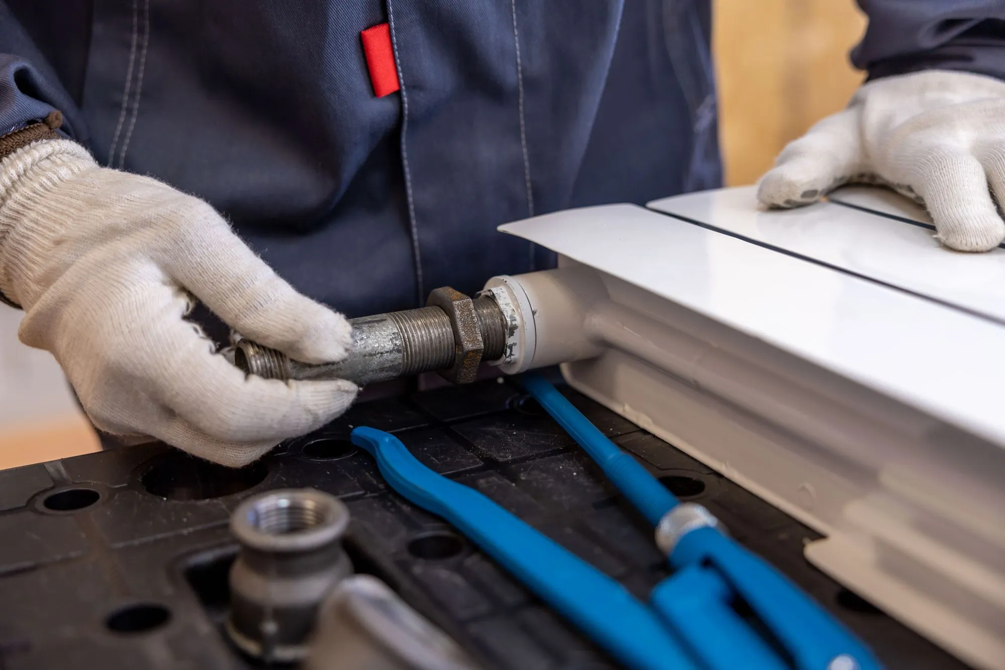 Gloved hands threading a fitting into a white panel radiator, with a pipe wrench on the bench