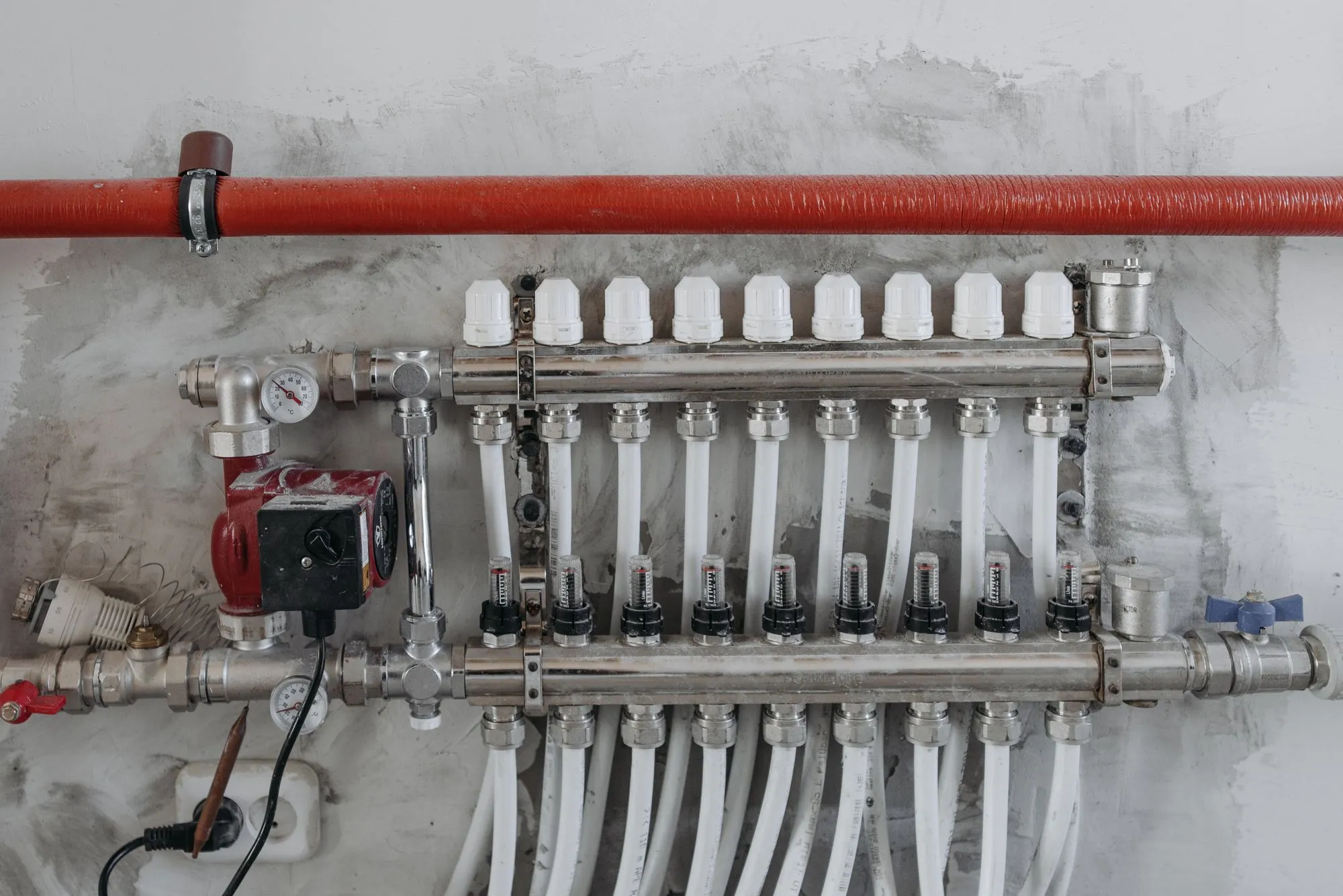 A radiant floor heating manifold with nine loops of white tubing and a red circulator pump